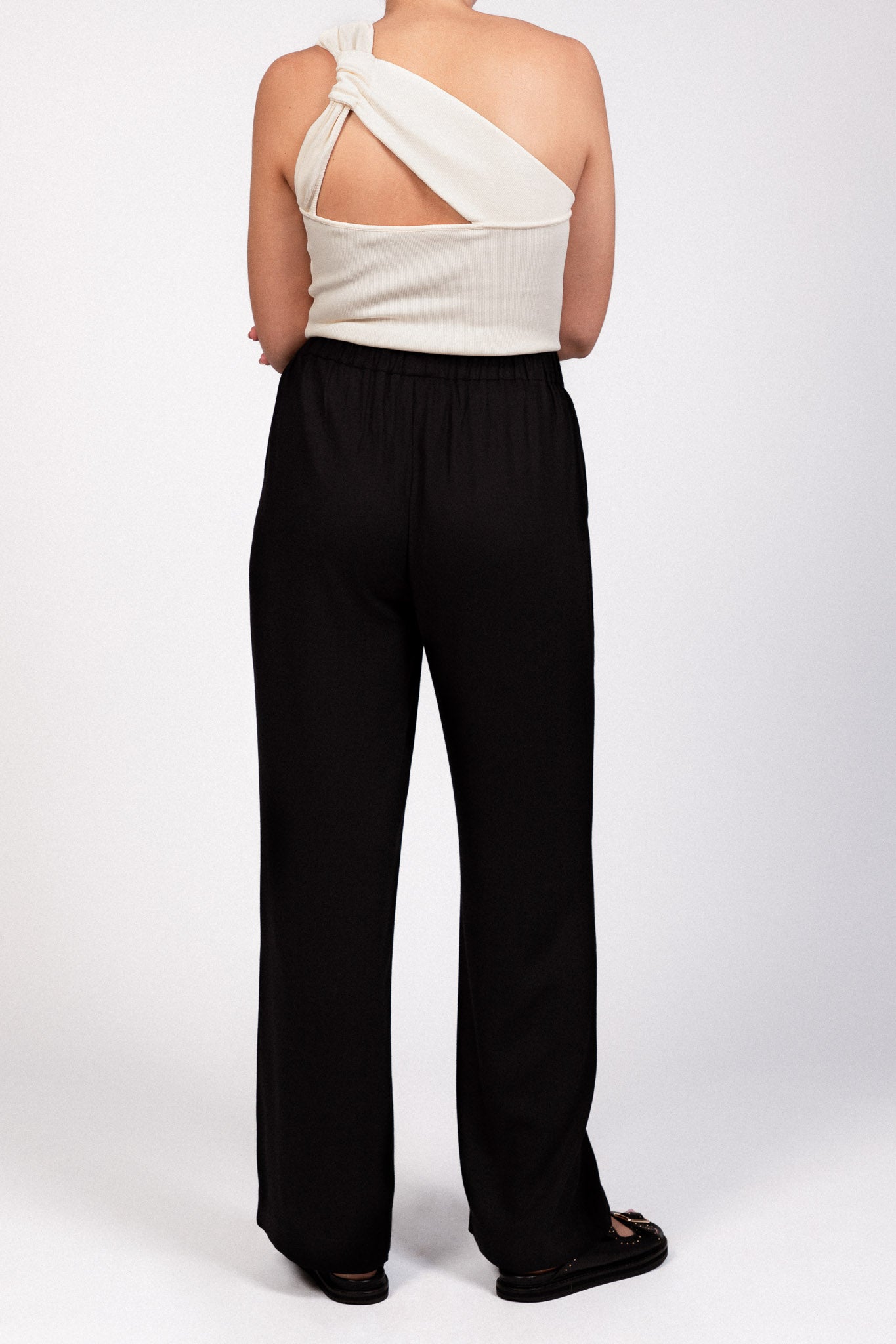 Pantalon noir fluide taille élastique MILO, coupe droite confortable, porté avec le top NOVA asymétrique écru, de dos - collection DILAA