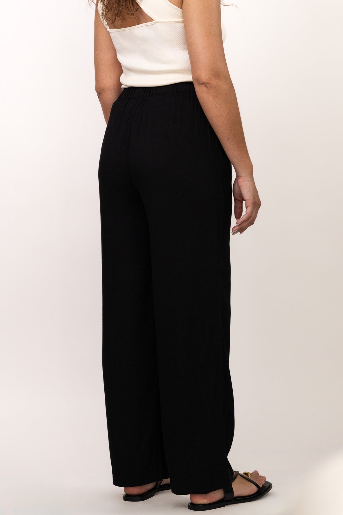 Pantalon noir fluide taille élastique MILO, coupe droite confortable, porté avec le top NOVA asymétrique écru, de dos - collection DILAA